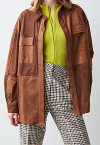 Brązowa, zamszowa, oversize koszula z dwoma kieszeniami na piersi, zestawiona z zielonym prążkowanym topem i czarno-białymi spodniami w kratę.