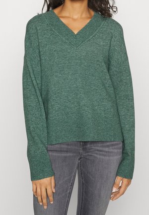 Femme portant un pull en maille vert col V avec poignets et ourlet côtelés, associé à un jean gris délavé, debout devant un fond uni.