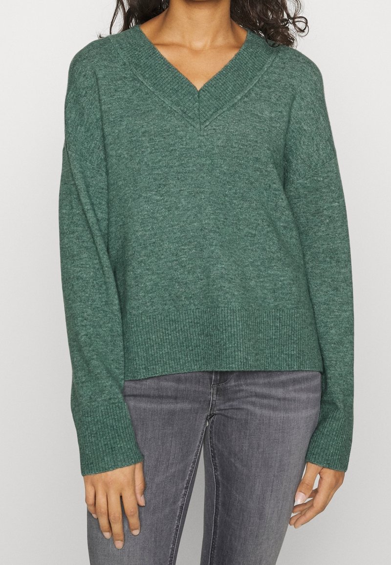 Femme portant un pull en maille vert col V avec poignets et ourlet côtelés, associé à un jean gris délavé, debout devant un fond uni.