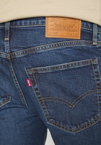 Tmavě modré džínové kalhoty s hnědou koženou záplatou na pasu, oranžovým prošíváním a červenou značkou Levi's na zadním kapse.