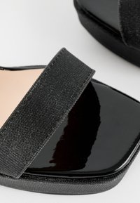 Černé sandály s lesklým patinovaným povrchem na podrážce a texturovaným černým popruhem. Čtvercový design špičky s polstrovanou stélkou.