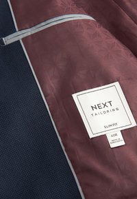 Veste de costume texturée bleu marine avec doublure bordeaux à motifs affichant une étiquette blanche indiquant "Next Tailoring Slim Fit 40R Made in Bangladesh".