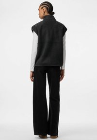 Melnas vilnas veste ar augstu apkakli, bezroku dizainu un teksturētu virsmu, valkāta virs baltas garām piedurknēm krekla un kombinēta ar melnām biksēm.