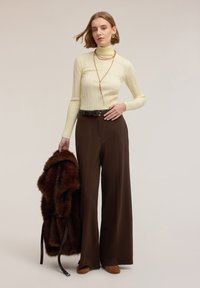 Kremowy prążkowany sweter z golfem, brązowe szerokie spodnie, brązowa kamizelka z futerka sztucznego przewiązana czarnym paskiem, zdobione brązowe buty.