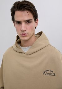 Jonge man met kort bruin haar, die een beige hoodie draagt met de tekst "Scalpers Time-Off Pizza, Pasta en Kleine Gerechten" op de borst, neutrale achtergrond.
