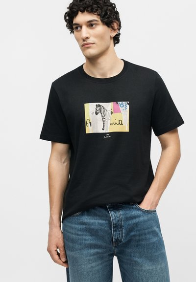 Νέος άνδρας με μπούκλες, που φοράει μαύρο μπλουζάκι με πολύχρωμο σχέδιο ζέβρας, συνδυασμένο με μπλε τζιν, στέκεται μπροστά σε απλό φόντο.