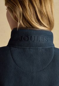 Persona con capelli biondi che indossa una giacca in pile blu scuro con collo alto ricamato con la parola "Joules" sulla schiena.