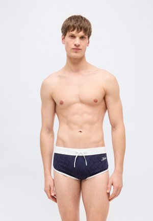 Jaunas vyras stovintis, dėvintis tamsiai mėlynas plaukimo glaudes su baltais apvadais ir raišteliu, prieš paprastą šviesiai pilką foną.