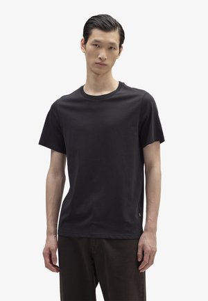 Jeune homme aux cheveux courts et foncés portant un t-shirt noir à manches courtes uni et un pantalon foncé, debout sur un fond blanc.