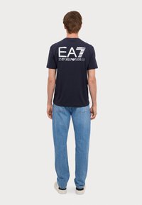 Tamsiai mėlynas medvilninis marškinėliai su dideliu baltu grafinės „EA7 EMPORIO ARMANI“ ant nugaros. Su šviesiai mėlynomis džinsinėmis kelnėmis.