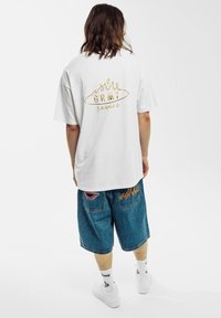 Camiseta blanca de gran tamaño con logo dorado en la parte trasera, combinada con pantalones cortos de mezclilla azules que presentan detalles de parches rojos y de llamas. Zapatillas blancas y calcetas.