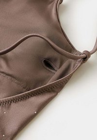 Gros plan d’un maillot de bain en tissu marron avec fines bretelles et un petit détail œillet sur fond blanc.