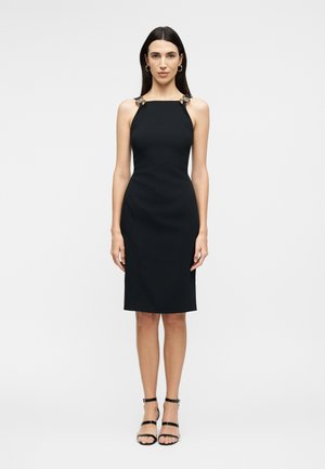 Γυναίκα με μακριά σκούρα μαλλιά, ντυμένη με εφαρμοστό μαύρο αμάνικο φόρεμα με διακοσμητικές τιράντες και μαύρα ψηλοτάκουνα σανδάλια με λουριά, όρθια.