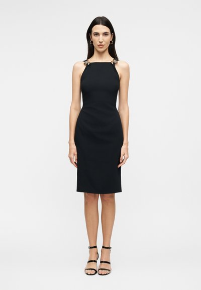 Femme aux longs cheveux foncés portant une robe noire ajustée sans manches avec des bretelles décoratives et des sandales à talons hauts à brides noires, debout.