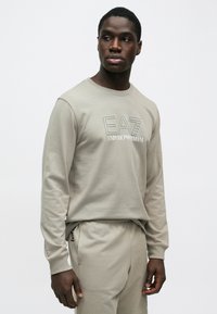 Egy férfi bézs Emporio Armani EA7 pulóvert és hozzáillő nadrágot visel, oldalra néz, egyszínű fehér háttér előtt.