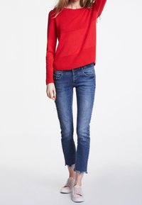 Frau in einem roten Langarmoberteil, blauen, eng anliegenden Jeans mit ausgefransten Säumen und hellrosa Sneakers, die vor einem weißen Hintergrund steht.