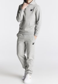 Grauer Kapuzenpullover und passende Jogginghosen aus weichem Stoff, mit einer vorderen Kängurutasche und schwarzen gestickten Logodetails. Weiße Sneaker runden den Look ab.