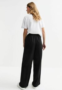 Witte t-shirt gecombineerd met zwarte broek met wijde pijpen en een elastische tailleband. Model draagt witte sneakers en toont een casual outfit.