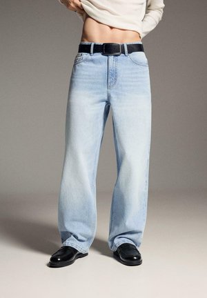 Jeans larghi color azzurro chiaro con cintura nera e scarpe nere, indossati da una persona che solleva una maglietta bianca per mostrare l'addome.