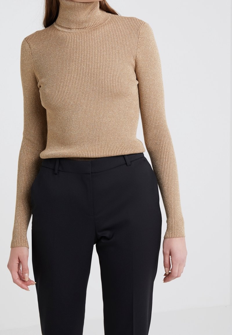 Femme portant un pull à col roulé côtelé beige et un pantalon noir sur mesure, debout devant un fond clair uni.