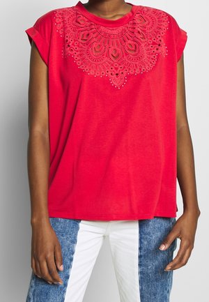 Personne portant un haut sans manches rouge vif avec des détails complexes en dentelle ajourée et perles au niveau du décolleté, associé à un jean patchwork bleu et blanc.