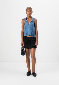 Denim brezrokaven top s koničastim ovratnikom in gumbom spredaj, v kombinaciji z črno plastično krilo z volančki in črnimi natikači.