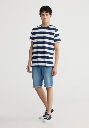 T-shirt a righe blu e bianche, maniche corte, scollatura tonda; abbinato a pantaloni shorts in denim azzurro e sneaker blu con suola bianca.