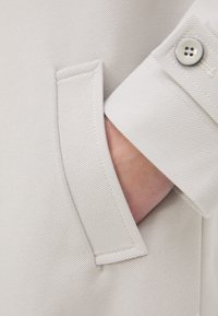 Lys beige stof med en tekstureret overflade, der har en sidelomme og en knapmanchet, som understreger rene linjer og et minimalistisk design.