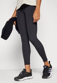 Leggings negros ceñidos con una textura suave. Par de zapatillas deportivas negras con tres franjas blancas y parte superior texturizada. Mano sosteniendo una toalla negra.