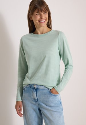 Femme souriante aux cheveux bruns portant une chemise à manches longues vert clair et un jean bleu clair, debout avec une main dans la poche.