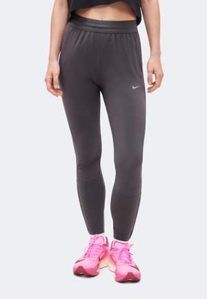 Femme portant des leggings Nike gris foncé et des chaussures de running Nike rose vif, debout devant un fond clair uni.