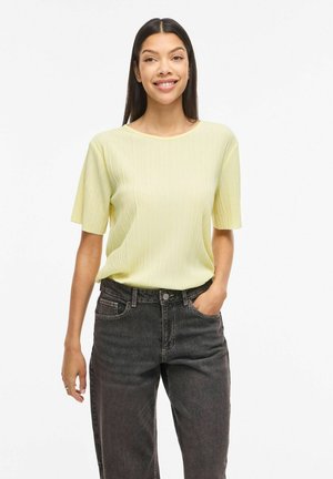 Usmievavá žena s dlhými tmavými vlasmi, oblečená v svetložltom krátkom rebrovanom topu a čiernych džínsach s vysokým pásom, s jednou rukou v vrecku.