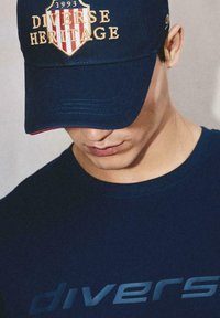Granatowa czapka baseballowa z złotą haftowaną aplikacją z napisem "Różnorodna Dziedzictwo" oraz rokiem "1993". Nosi się ją z dopasowaną granatową koszulką, co podkreśla minimalistyczny design.
