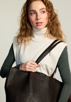 Young woman with curly hair wearing a white sleeveless turtleneck over a dark long-sleeve shirt, holding a large dark brown leather tote bag.