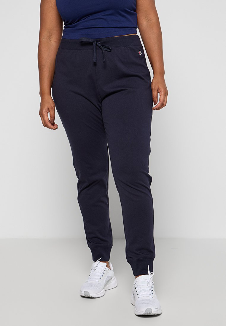 Marinefarbene Sweatpants mit einem Kordelzug, gerippten Bündchen, weichem Stoff und einem kleinen Logo an der Hüfte, kombiniert mit weißen Sportschuhen.
