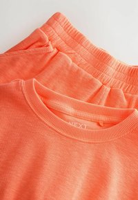 Due capi a coste di un vivace arancione, comprendenti una maglietta a girocollo e un pantalone con fascia elastica in vita, che mostrano un tessuto leggero e testurizzato.