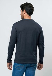 Homme aux cheveux foncés portant une chemise noire à manches longues et un jean bleu, debout dos à la caméra sur un fond uni.