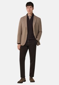 Blazer marrone in lana con colletto a revers, indossato sopra un maglione nero con zip e una camicia chiara, abbinato a pantaloni scuri e scarpe marroni.