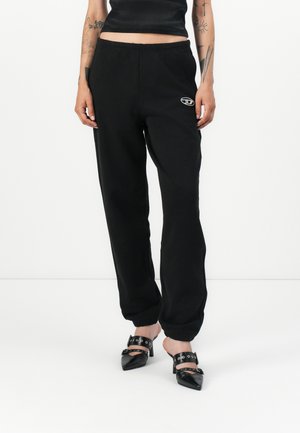 Black jogger pants with elastic waistband and cuffs, featuring a small logo. Soft material with a relaxed fit. Worn with pointed black heels.