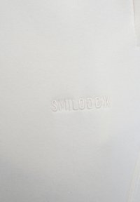 Valkoinen collegepaita, jossa on brodeerattu brändinimi "SMILODOX" hienovaraisella, vastaavalla langalla. Pehmeä rakenne ja istuva muotoilu.