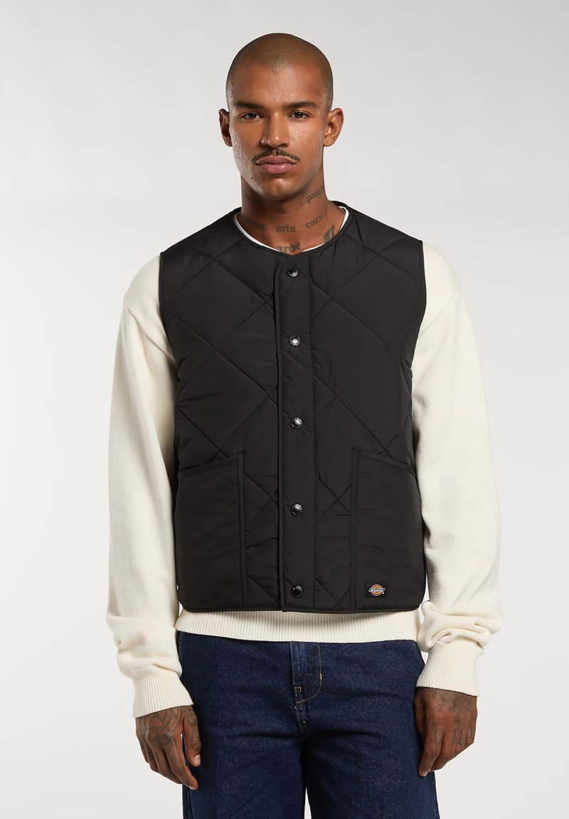Chaleco negro acolchado con botones a presión, dos bolsillos frontales y un acento de logo. Llevado sobre un suéter crema, con cuello redondo y mangas largas.