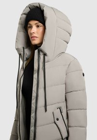 Chaqueta acolchada gris con una gran capucha, cordones ajustables negros, cremallera frontal y un bolsillo lateral con botón a presión. Gorro de punto negro adjunto.