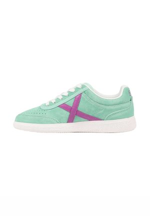 Zapatilla deportiva de ante verde claro con suela y cordones blancos, con un gran diseño de X púrpura en el lateral, vista desde el lado exterior.