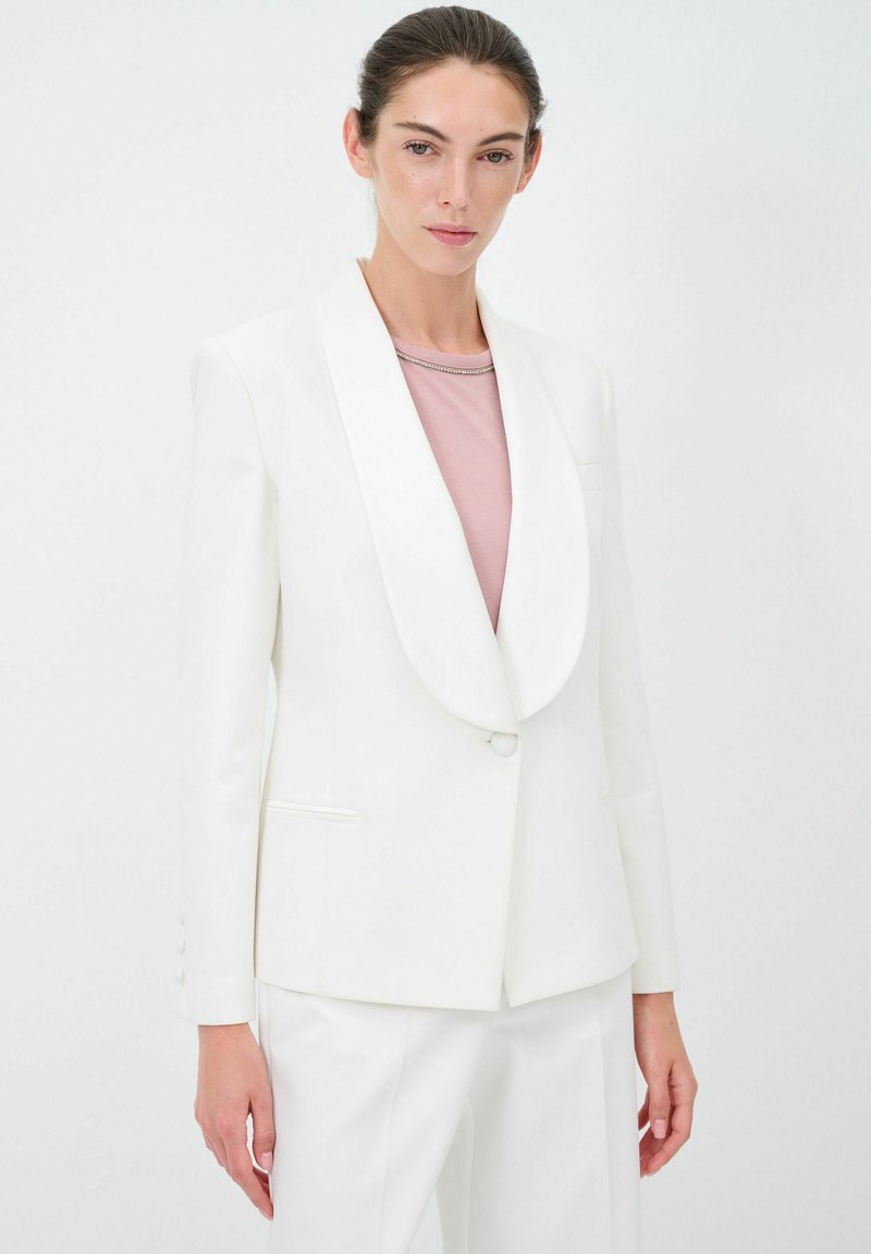 Blazer bianco con rever a scialle, chiusura a un bottone e due tasche frontali. Indossato sopra una camicia rosa chiaro con un sottile dettaglio sul collo.