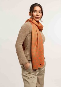 Écharpe orange en tissu texturé, ornée d'un motif de lèvres répétitif, drapée autour du cou, associée à un pull léger marron et à un pantalon beige.