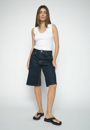 Ung kvinde iført hvid ærmeløs top, mørke knælange denimshorts og sorte platåsandaler stående mod en ensfarvet baggrund.