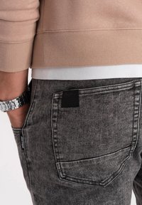 Grå denimjeans med en strukturerad finish, som har en fyrkantig svart lap på bakfickan och en kontrasterande vit midja under en beige sweatshirt.