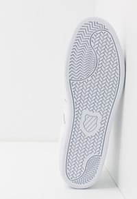 Vit sneakersula med herringbone-mönster, prydd med ett sköldemblem och en slät strukturerad yta.