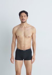 Boxeri negri dintr-un material elastic, cu un talie moale, purtând numele brandului. Modelul stă cu ochii închiși pe un fundal alb.