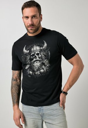 Mann trägt schwarzes T-Shirt mit detailliertem Totenkopf-Viking-Helm-Design, helle Jeans und tätowierten rechten Arm, steht vor einfachem Hintergrund.